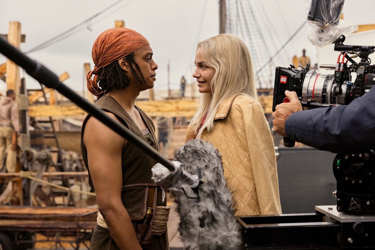 Two costumed actors face each other on a shipyard set while a boom microphone and a large cinema camera frame the shot