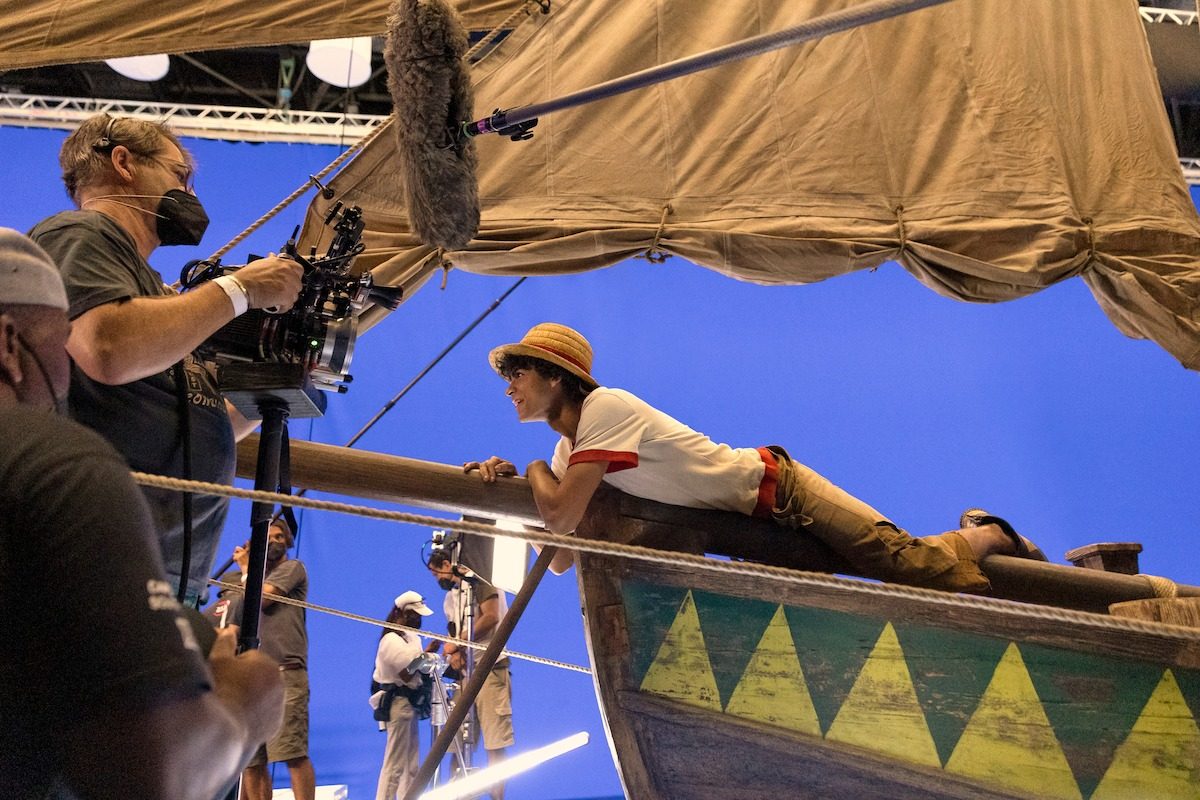 A costumed actor in a straw hat lies along the bow rail of a ship set against a blue screen while an operator films handheld